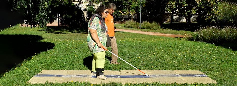 Test du dispositif CAU-mobility par une personne aveugle Test du dispositif CAU-mobility par une personne aveugle, le cheminement est placé sur une pelouse, la personne circule de gauche à droite, sa canne détecte les reliefs qui guident son parcours