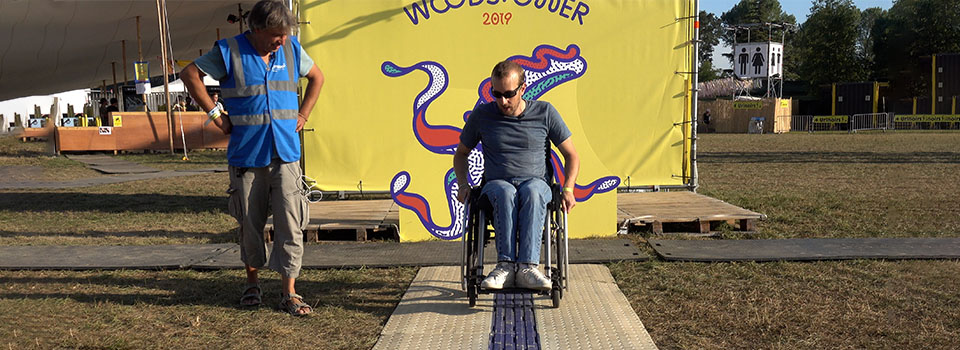 Test du CAU-mobility au festival Woostower Une personne en fauteuil roulant test le CAU-mobility installé sur l’herbe au festival Woodstower dont l’affiche se trouve en fond. Différence de couleur et de texture entre la CAU-mobility (beige latéralement et bleu au centre) et les plaques de roulages (toutes grises). Les modules du CAU-mobility sont ajusté, avec des côtés en pente accessible aux fauteuils contrairement aux plaques de roulage (solution par défaut et inadaptée).
