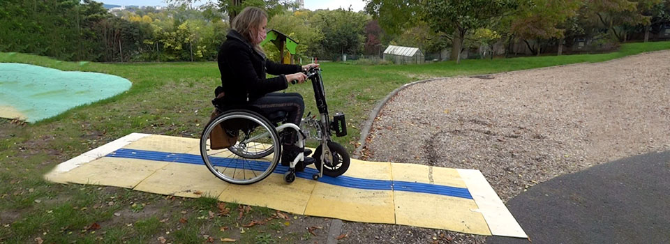 CAU-mobility convient aux fauteuils roulants adaptés sur terrains accidentés Personne en fauteuil roulant manuel augmentée d’une roue électrique roule sur le CAU-mobility