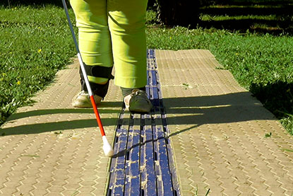 Test canne d’une personne aveugle Une personne aveugle marche avec sa canne sur le CAU-mobility, installé sur l’herbe. La photo montre en gros plan le toucher de canne sur les cannelures au milieu du cheminement